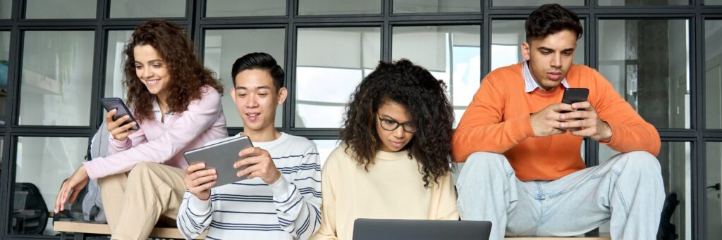 “Four young adults sit together indoors, each focused on a different digital device. One woman smiles at her phone, a man next to her looks at a tablet, another woman works on a laptop, and a man on the end concentrates on his phone. They are seated in front of large glass-paneled windows