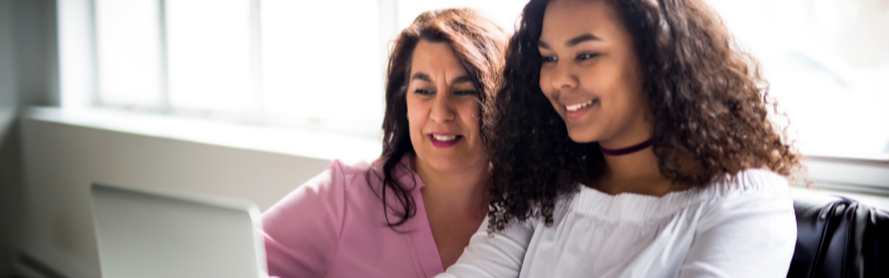 A woman and a teenage girl sit together looking at a laptop screen. Both are smiling, and the girl appears to be leading the activity while the woman looks on supportively. They are seated by large windows with natural light coming in.