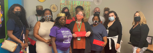A group of staff members stand together indoors, all wearing face masks. Some people wear gold paper crowns, and several hold red envelopes. They are posed in front of a decorated wall with colorful hanging streamers and paper decorations, smiling for the group photo.