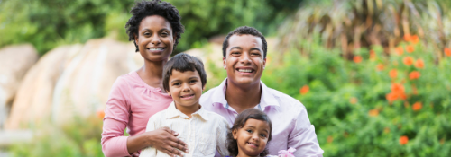A smiling family of four poses outdoors in a garden setting. Two adults stand behind their two young children, with greenery and flowers in the background.