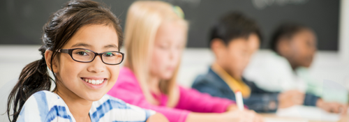 A smiling young girl wearing glasses sits at a classroom desk, looking toward the camera. Behind her, other children are focused on writing or schoolwork, with a chalkboard in the background.