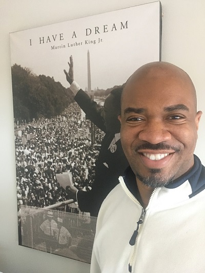 Dereck Dean smiling for a picture infront of a poster of Martin Luther King Jr. I have a Dream speech.