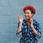 Young African American lady with bright red hair, smiling and pointing off into the distance standing against a blue brick wall.