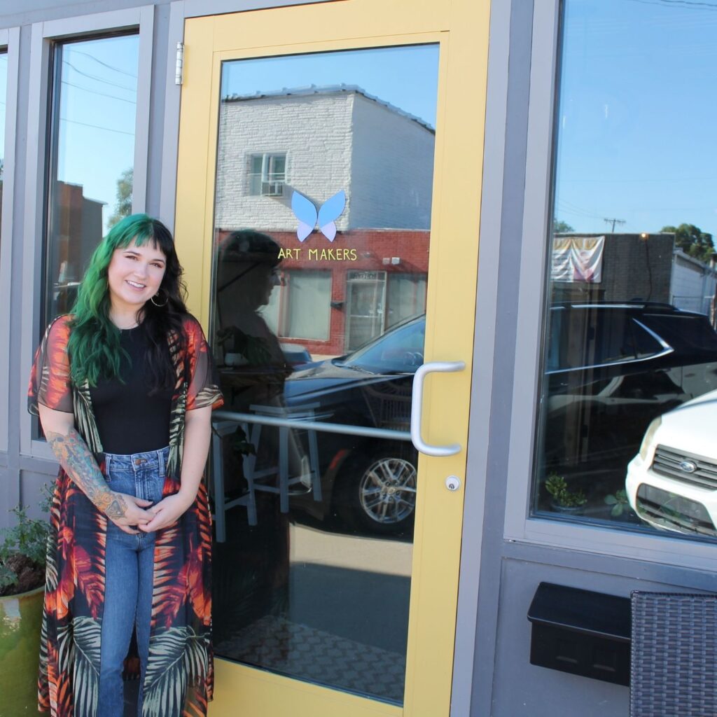A woman stands outside of yellow business doors with the Art Makers logo.